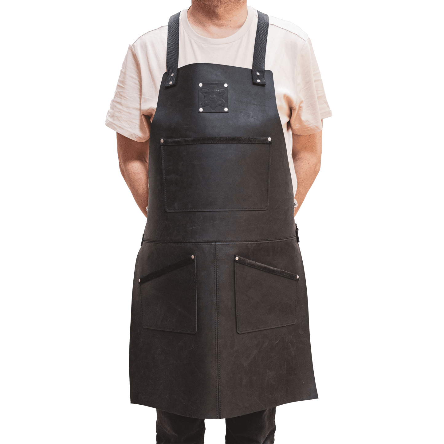 A carpenter wearing a dark leather apron from The Durham Leatherworker over a casual white t-shirt, standing with his arms subtly to his sides, against a black background.