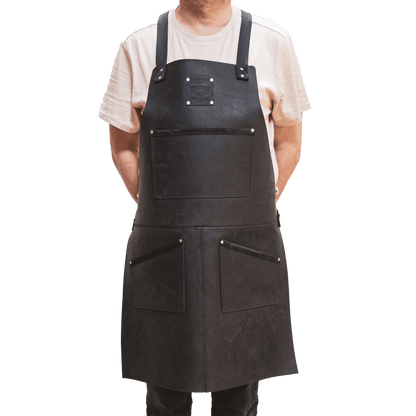 A carpenter wearing a dark leather apron from The Durham Leatherworker over a casual white t-shirt, standing with his arms subtly to his sides, against a black background.