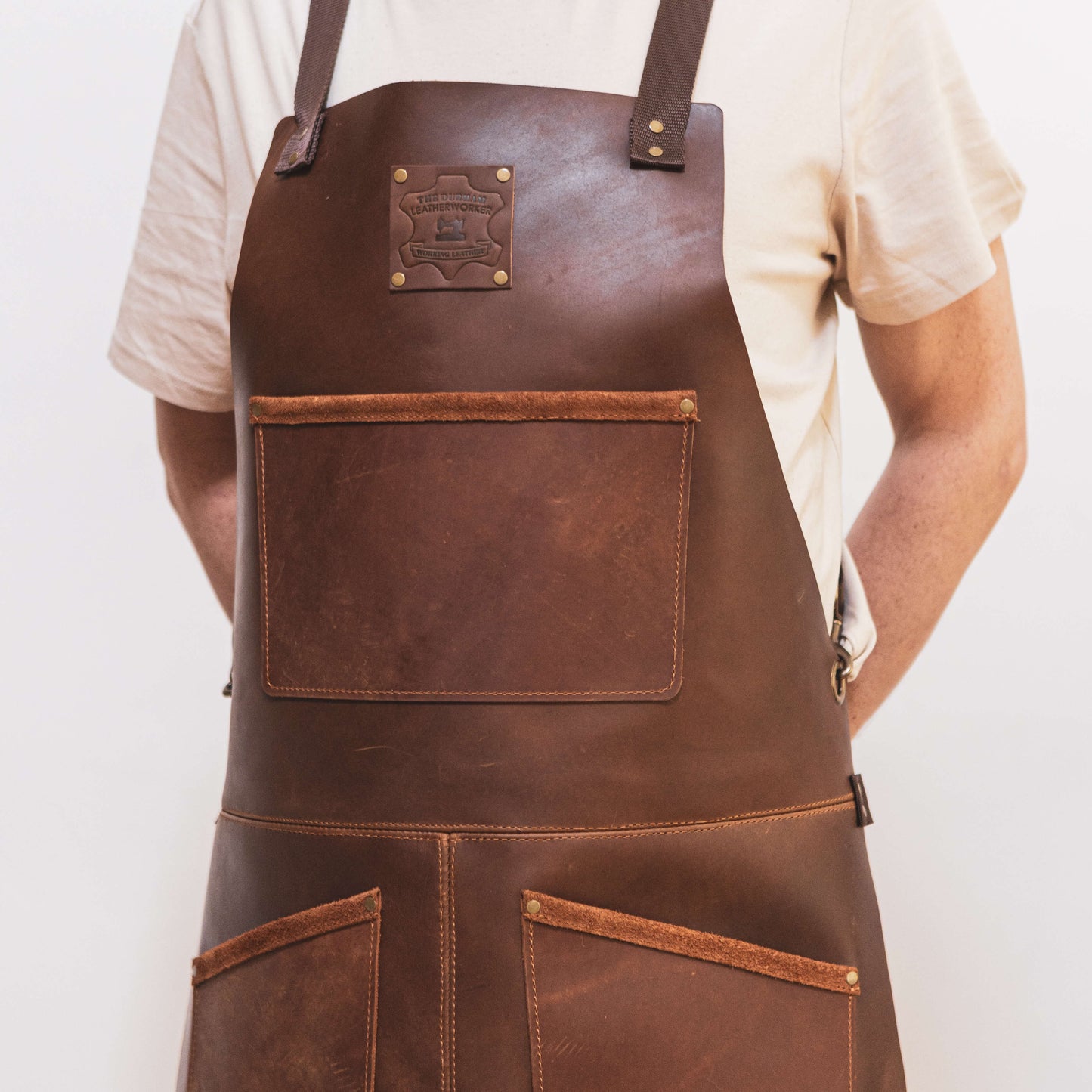 A tradesman wearing a white shirt and a dark brown leather apron from The Durham Leatherworker, with pockets and a logo patch, standing against a plain background. Only the torso is visible.