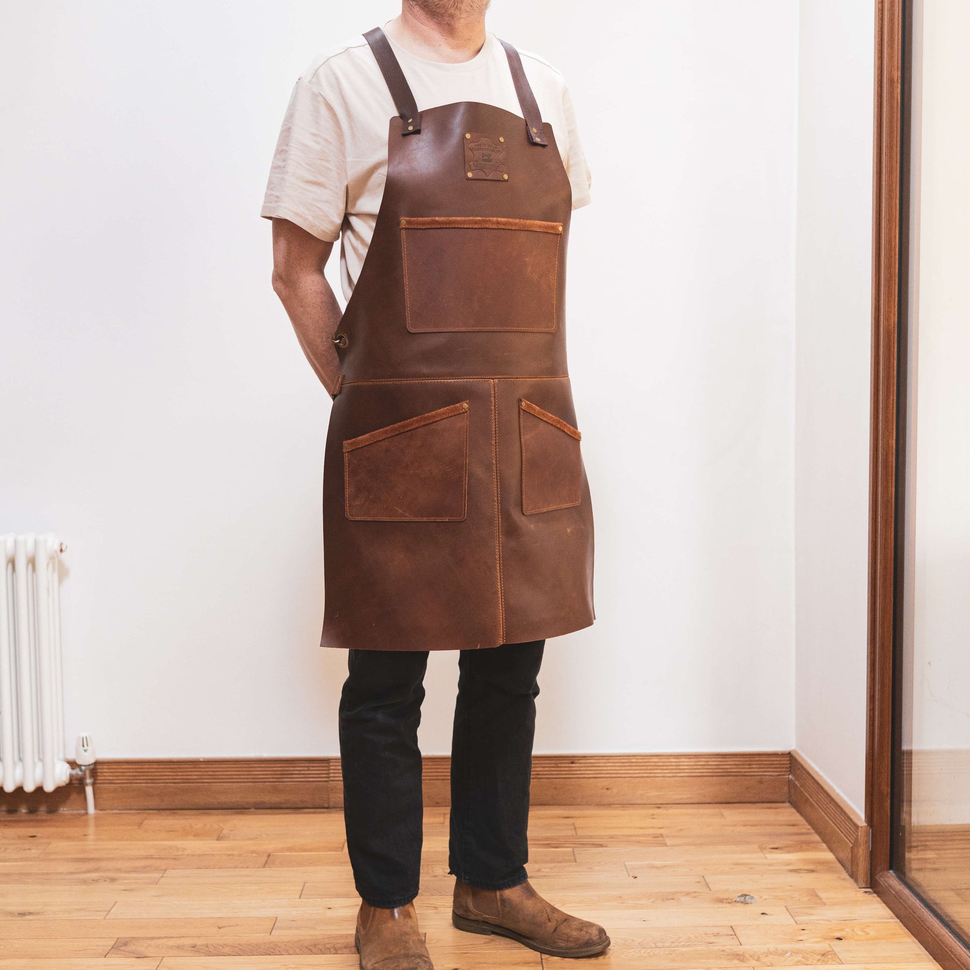 A carpenter wearing The Durham Leatherworker leather apron stands in a room with wooden flooring, facing a mirror. The apron has multiple pockets, and the person is dressed casually in a white t-shirt and dark jeans.