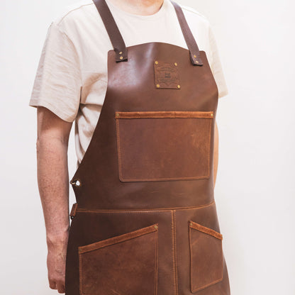 A carpenter wearing a brown leather apron from The Durham Leatherworker with pockets and adjustable straps stands against a plain white background, visible from the chest down.