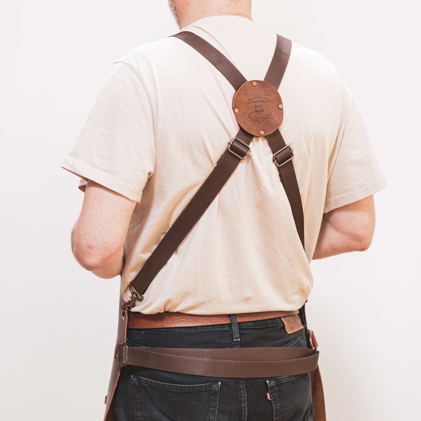 Rear view of a carpenter wearing a beige t-shirt, dark brown leather suspender toolbelt from The Durham Leatherworker, and a belt, featuring a brass round centerpiece on the suspenders against a light background.