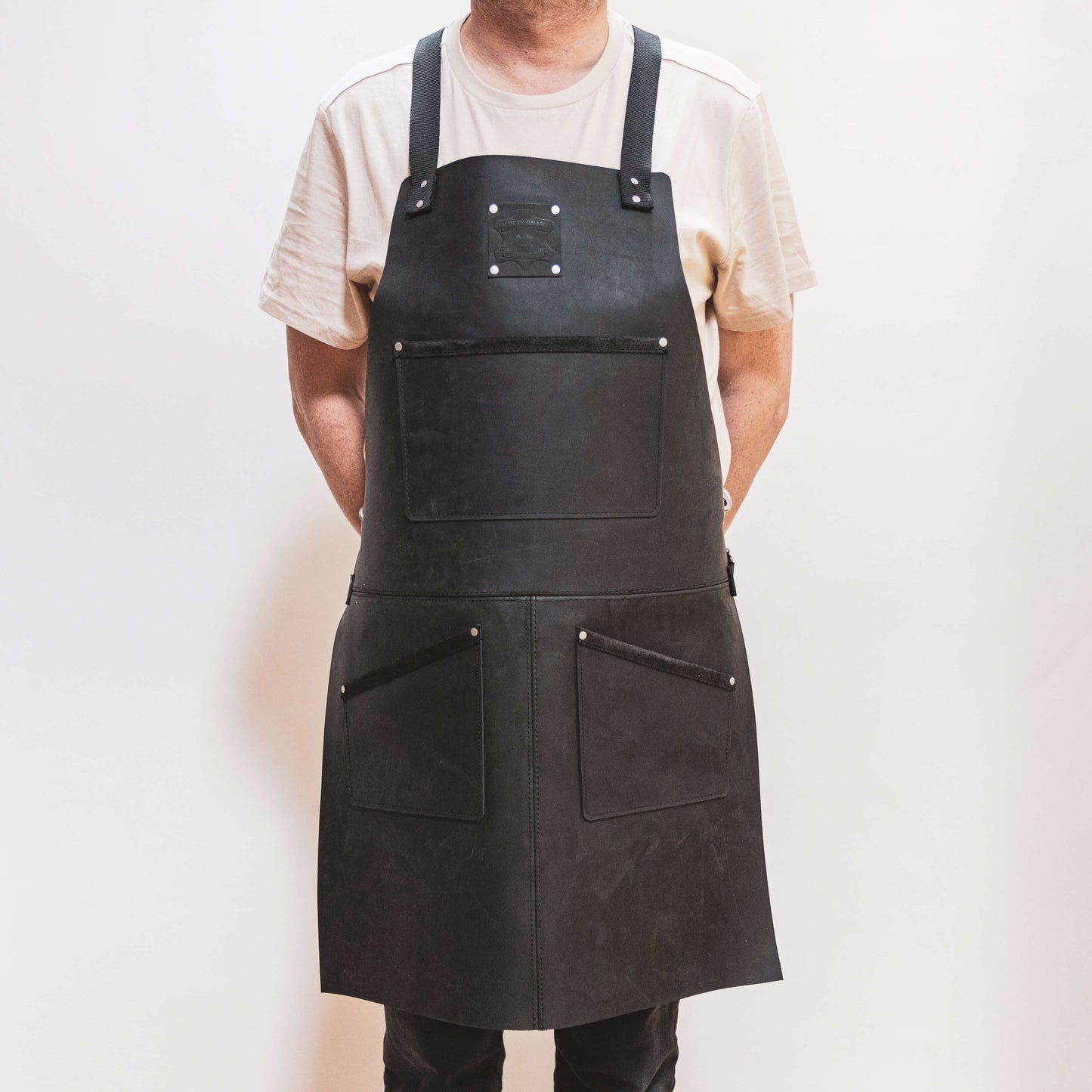 A carpenter wearing a white t-shirt and The Durham Leatherworker dark leather apron stands against a light background, with the apron featuring pockets and metallic clasps.