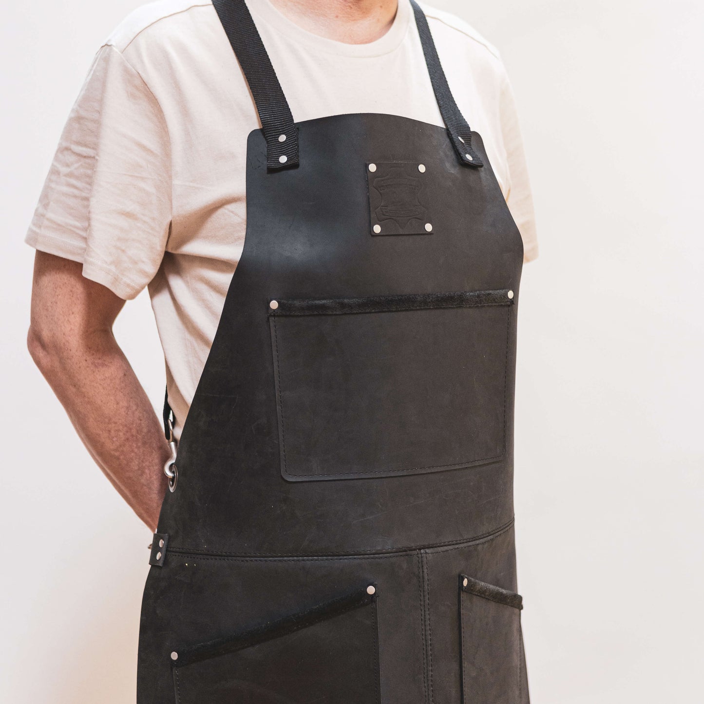 A joiner wearing a dark leather apron with straps over a beige t-shirt, against a light background. The Durham Leatherworker apron covers from chest to knees, displaying multiple pockets and silver rivets.