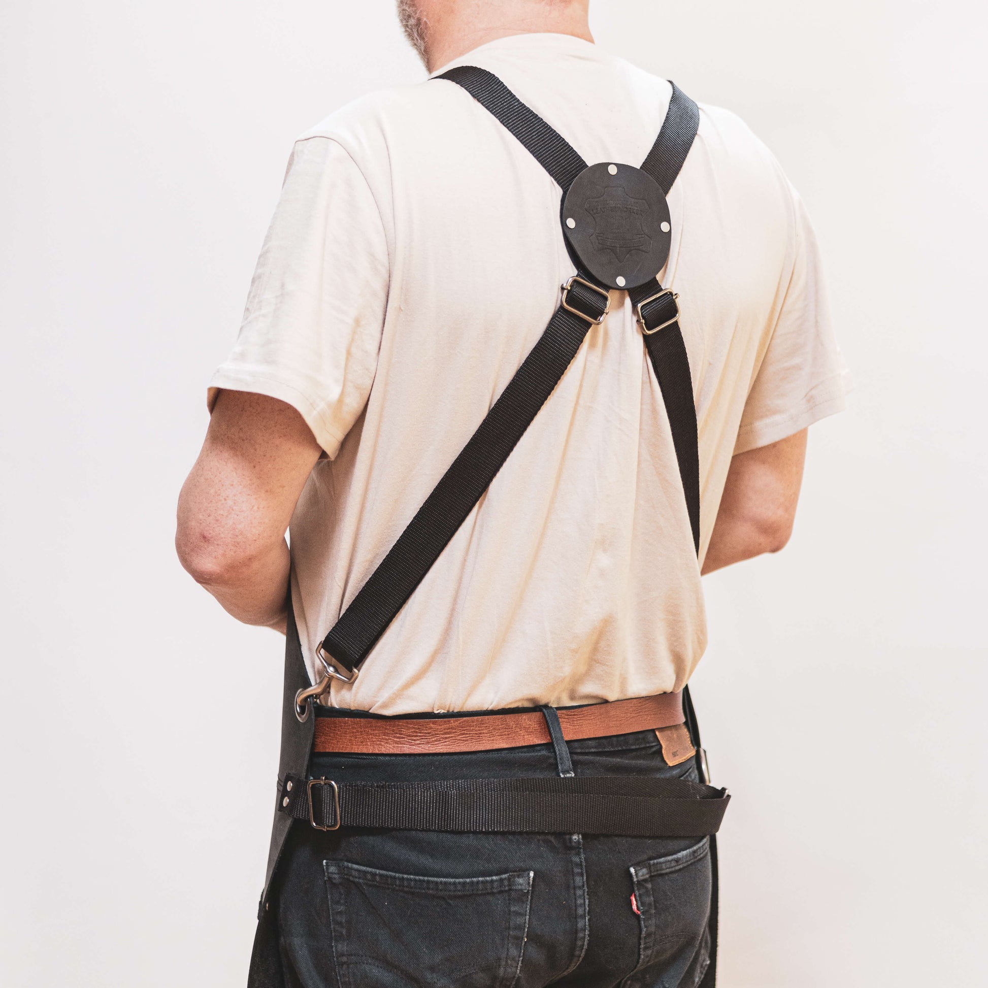 A man viewed from behind wearing a cream shirt and denim jeans, with a black crossbody bag designed by The Durham Leatherworker for tradesmen and a brown leather belt. The focus is on the way he carries the bag.