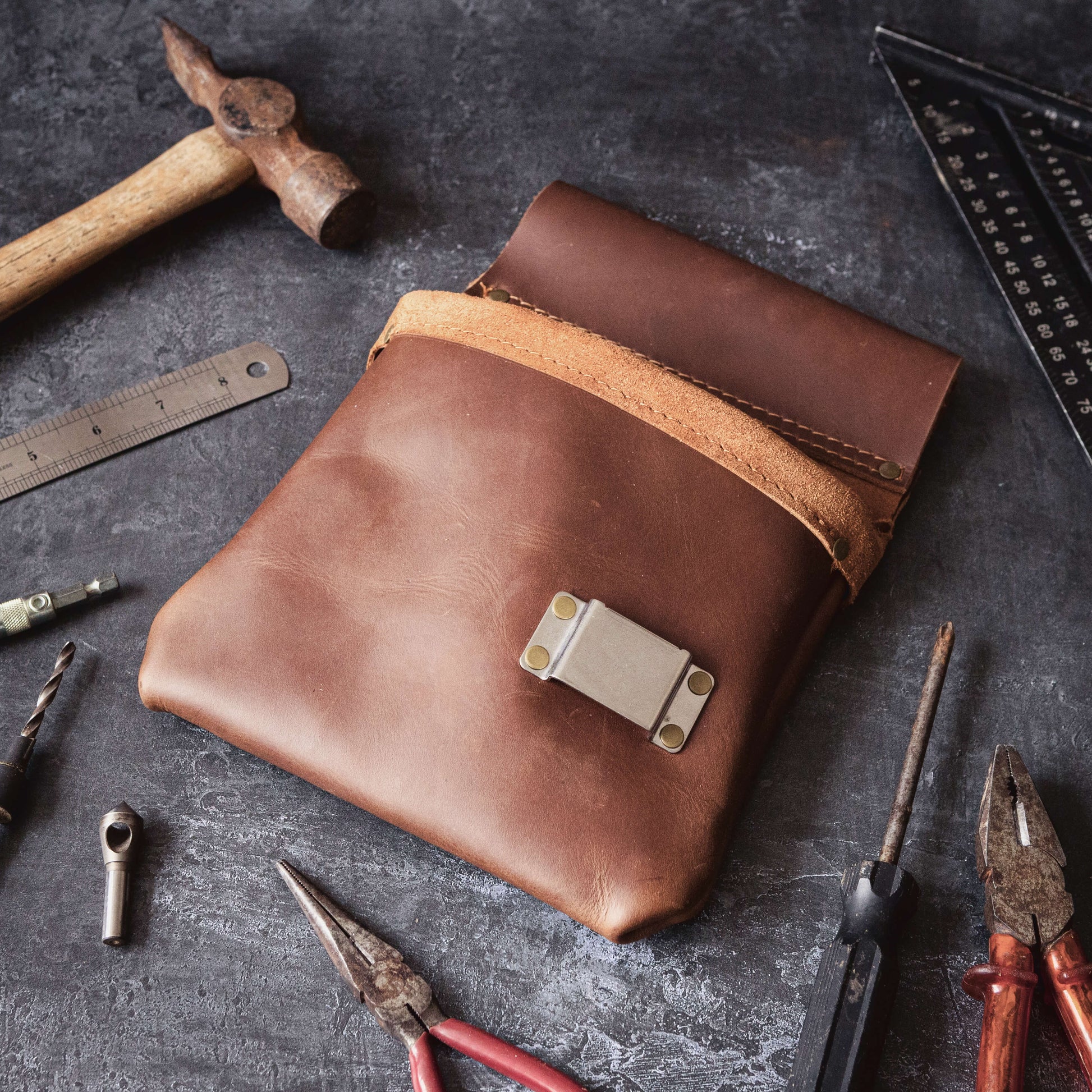 A Belt Bag 3 from The Durham Leatherworker lies amidst various tools like hammers, pliers, and rulers on a textured grey surface, representing a carpenter's workspace.