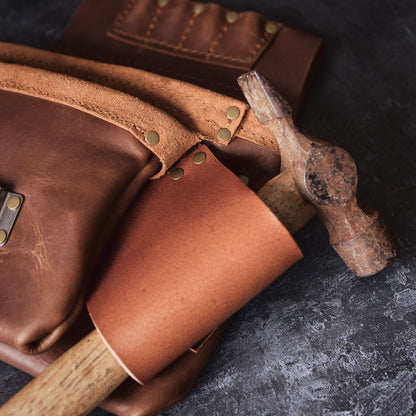 A close-up of leather crafting tools, including a rustic hammer and brown leather pieces with brass rivets, arranged on a textured dark surface in The Durham Leatherworker's Belt Bag 1.
