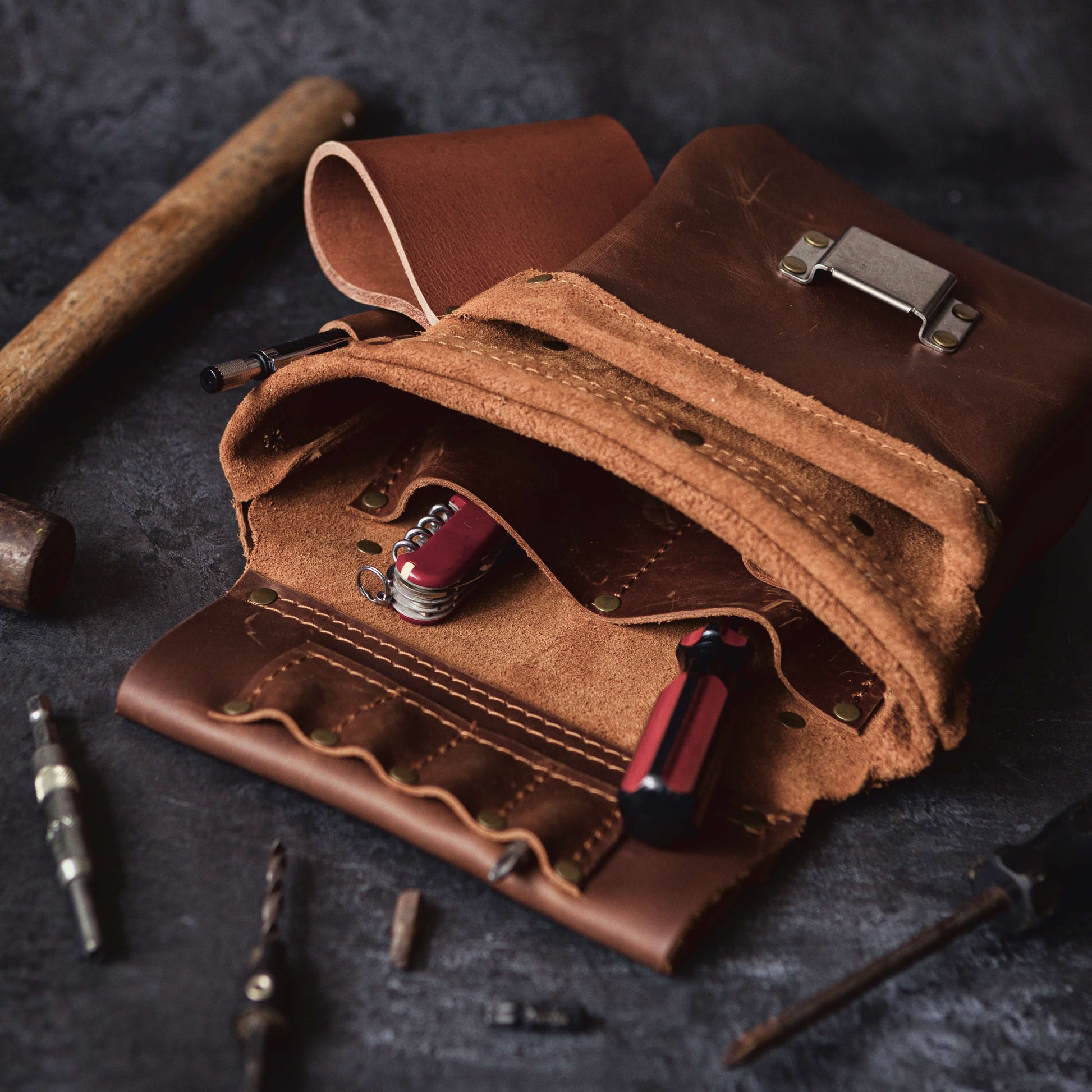 A rustic leather Belt Bag 1 by The Durham Leatherworker, partially open, displays various small tools like screwdrivers inside, with two pocket flashlights also visible. The roll rests on a dark, textured surface alongside additional woodwork tools.