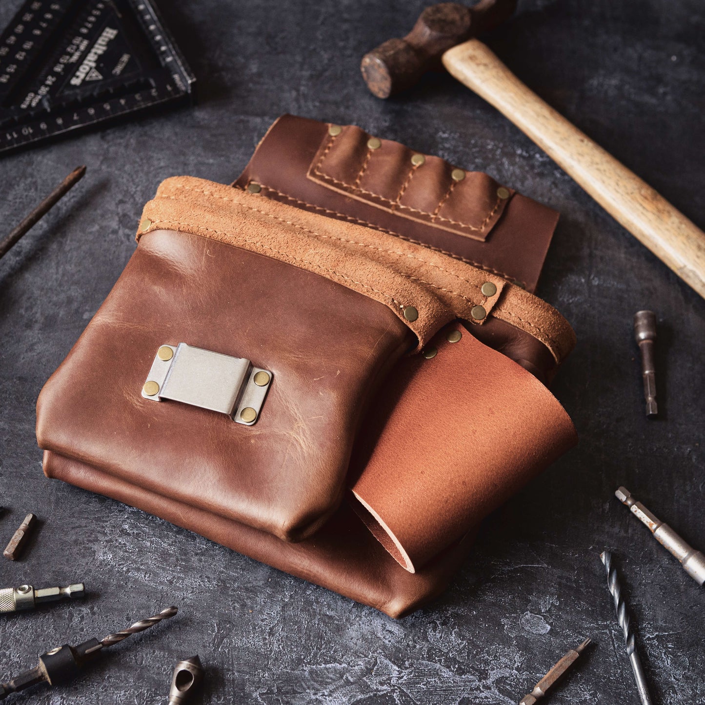 A collection of handcrafted leather goods, including a Belt Bag 1 by The Durham Leatherworker, tool roll, and journal cover, displayed alongside carpenter's metal tools on a dark, textured surface.