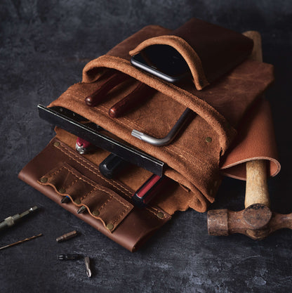 An open carpenter's leather tool roll containing various tools, including a hammer and screwdrivers, with a smartphone also tucked inside, displayed on a dark textured surface.