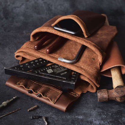 A still life of a rustic leather tool roll from The Durham Leatherworker containing a hammer, various screwdrivers, and a folding ruler, accompanied by a smartphone, set against a dark, textured background designed for woodwork.