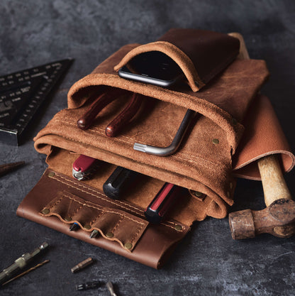 A close-up of a well-organized Belt Bag 2 from The Durham Leatherworker containing various tools like screwdrivers and a hammer, alongside a smartphone, all placed on a dark, textured surface.